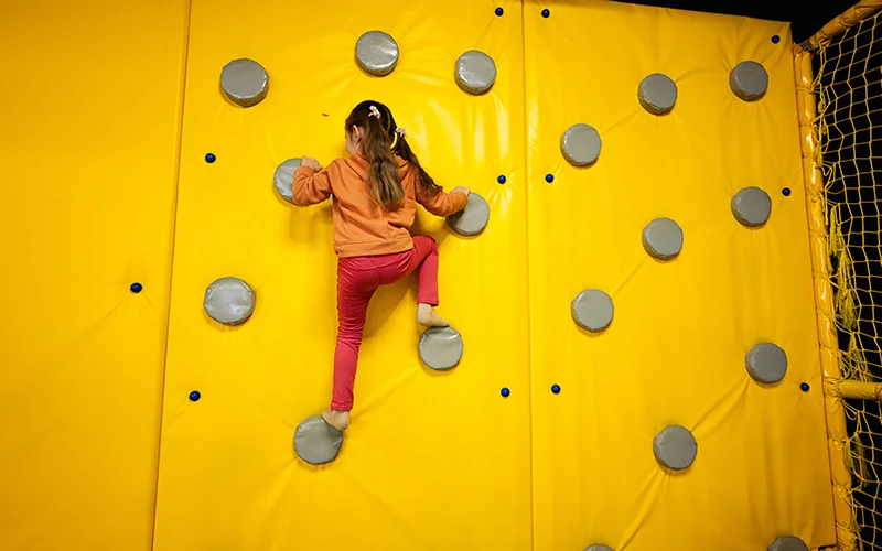 Child climbing a soft climbing wall in an indoor playground adventure area.