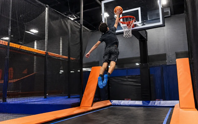Boy performing an airborne dunk on a trampoline at an indoor trampoline park, showcasing athletic skill and fun