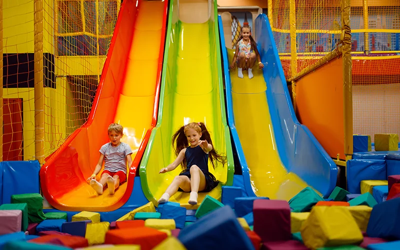 Three children having fun sliding down colorful indoor playground slides onto a soft foam pit