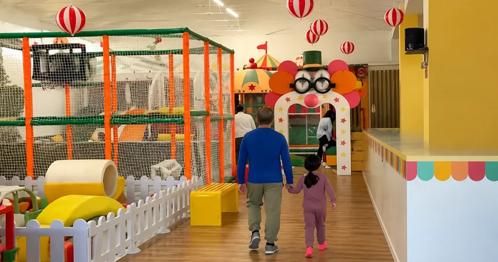 Parent and child walking in an Italy circus-themed indoor playground, showing colorful play structures, safety fencing, and a circus-themed entrance arch.
