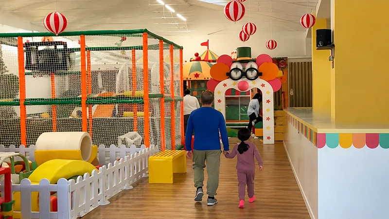 Parent and child walking in an Italy circus-themed indoor playground, showing colorful play structures, safety fencing, and a circus-themed entrance arch.